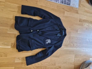 Svart kavaj från Polo Ralph Lauren - Svart kavaj från Polo Ralph Lauren med klassisk passform och två framfickor. Snyggt broderat PRL-emblem på bröstet och stilrena knappar framtill. Tillverkad i mjuk bomull som känns skön mot huden och elastiskt i passform.Perfekt för dig som vill ha en clean och tidlös look. Använd fåtal gånger. Nästan som ny.