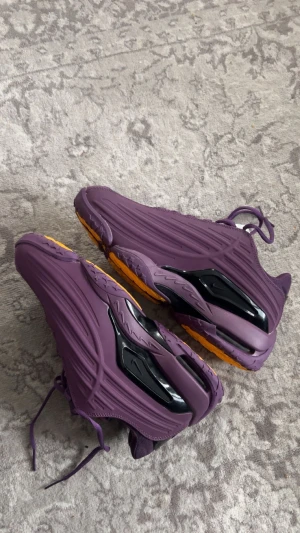 Lila Nike sneakers med gul sula - Säljer ett par unika lila Nike sneakers med coola svarta detaljer och en kontrasterande gul sula med stjärnmönster. Skorna har snörning och en futuristisk design med platt sula och rund tå. Perfekt för dig som vill sticka ut med färg och stil.