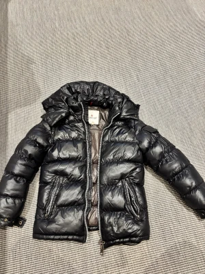 Svart Moncler pufferjacka med huva - Säljer en svart pufferjacka från Moncler med glansig finish och huva. Jackan har två dragkedjefickor, quiltad design och coolt Moncler-märke på ärmen. Insidan har ett serietidningsmotiv och en innerficka med dragkedja. Perfekt för kalla dagar!