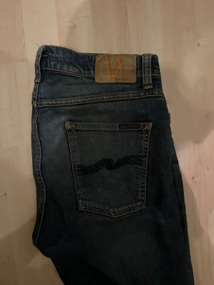 Nudie Jeans  - Säljer ett par Nudie Jeans Lean Dean i färgen Indigo Shades. De är tillverkade av 99% ekologisk bomull och har en klassisk femficksdesign med orangea sömmar och Nudie-logga på bakfickan. Jeansen har en slim passform och raka ben med snygg tvätt.