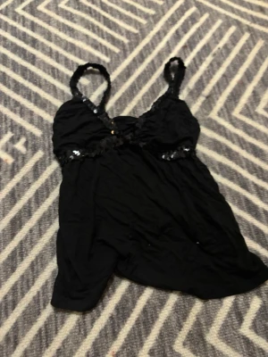 Svart linnetopp med paljettdetaljer - Säljer en svart linnetopp med smala axelband dekorerade med svarta paljetter. Toppen har en mjukt rynkad byst och ett lätt, flödande tyg som ger en skön känsla. Märke eller storlek står inte, men skulle säga att det är XS/S, materialet är väldigt stretchigt.