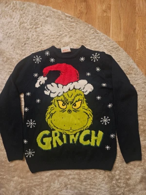 Svart Grinch jultröja från Dr. Seuss - Rolig svart stickad tröja med stor Grinch-tryck i grönt och gult, röd tomteluva och vita snöflingor. Perfekt för julen och har rund hals samt långa ärmar. Märkt med The Grinch från Dr. Seuss.