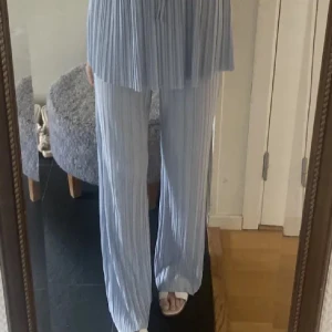 Plisserade ljusblå byxor från Zara - Säljer ett par plisserade ljusblå byxor från Zara med loose passform och elastisk midja. Byxorna har ett snyggt ribbat tyg och är tillverkade i ett mjukt syntetmaterial som ger en skön känsla. Perfekta för dig som gillar en chill och trendig look.                  Tillhörande matchande linne i samma plisserade material med smala axelband och lätt v-ringning medföljer. Perfekt set för dig som gillar en chill och trendig look.