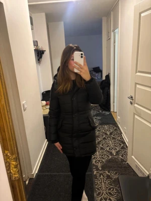 Svart pufferjacka från GANT, S - Säljer en svart pufferjacka från GANT i storlek S. Jackan har huva, dragkedja och knappar framtill samt två fickor med lock. Midjan är justerbar! Den är quiltad och har en längre modell som håller dig varm under kalla dagar. Perfekt för dig som gillar stilren och funktionell design.