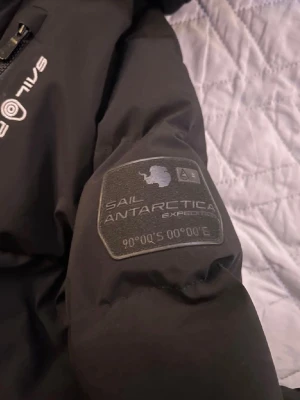Svart pufferjacka från Sail Racing - Svart pufferjacka från Sail Racing med två bröstfickor med dragkedja och hög krage. Jackan har snygga Sail Antarctica Expedition-patch på ärmen och är designad för att klara kalla dagar. Perfekt för dig som gillar stil och funktion.