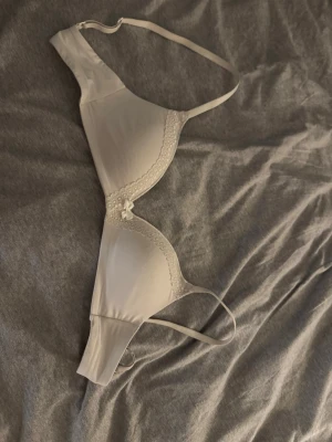 Vit spetsdetaljerad bh - Säljer en vit bh med fina spetsdetaljer längs kanten och en liten rosett framtill. Bh:n har justerbara axelband och slät design som ger en enkel och stilren look.  Inte Push-Up Kan inte ta av banden eller ta ur ”paden” Inga bågar