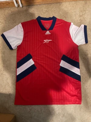Röd Adidas Arsenal fotbollströja - Röd fotbollströja från Adidas med vita ärmar och mörkblå detaljer. Tröjan har Arsenal-logga på bröstet och klassiska Adidas-ränder i vitt och blått på sidorna. Perfekt för dig som älskar fotboll och vill visa ditt stöd för Arsenal.