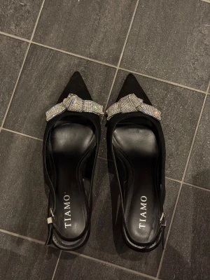 Svarta pumps med glittrig rosett från Tiamo - Säljer svarta klackskor från Tiamo med spetsig tå och låg klack (5cm). Glittrig rosett framtill och en öppen häl med justerbart spänne. Materialet är mocka och insidan är i skinn. Mjuk sula och lätta att gå i. Perfekt för dig som gillar att sticka ut med detaljer. Nypris i butik: 799