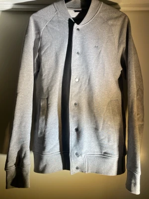 Grå J.Lindeberg cardigan kofta - En grå cardigan från J.Lindeberg med en minimalistisk färg med en grym passform. Den har en knapprad, sidofickor och ribbade manschetter på ärmar och fåll samt en ribbad krage. Riktigt snygg, klassisk och perfekt till alla kläder. 