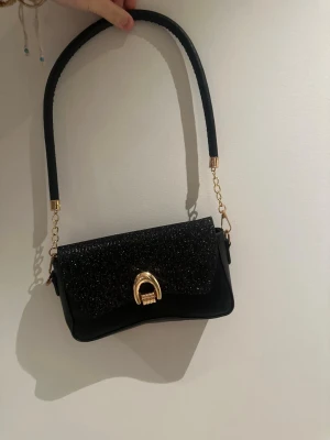 Svart glittrig axelväska med gulddetalj - Snygg svart axelväska med glittrigt lock och guldfärgat spänne framtill. Perfekt storlek för mobil, plånbok och småsaker. 