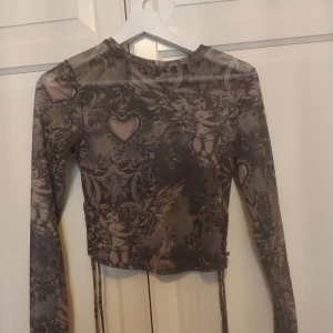 Grå mesh topp med änglamönster FB Sister - Säljer en grå mesh topp från FB Sister i storlek S. Toppen har lång ärm, snörning på sidorna och ett detaljerat mönster med änglar, hjärtan och ornament. Croppad passform och transparent mesh-material som ger en edgy vibe.