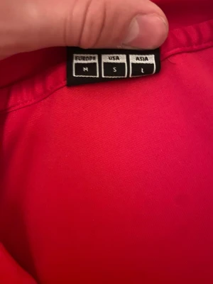 Röd Hummel t-shirt med vita detaljer - Säljer en röd Hummel t-shirt i storlek M med vita detaljer på axlarna och runt halsen. Tröjan har korta ärmar och är gjord i ett lätt syntetmaterial som passar perfekt för träning. Klassisk Hummel-logga på bröstet och snygga vita pilar på axlarna.