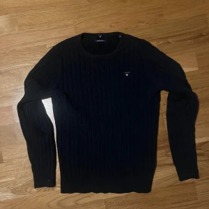 Mörkblå kabelstickad tröja GANT - Snygg mörkblå kabelstickad tröja från GANT i stretchig bomull. Tröjan har rund hals, lång ärm och diskret broderad logga på bröstet. Perfekt för dig som gillar klassisk och stilren look med lite preppy vibe.