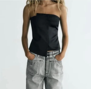 Svart asymmetrisk tubtopp från Zara - Säljer en svart tubtopp från Zara med cool asymmetrisk design och stilren look. Toppen är axelbandslös och har en snygg omlott-detalj framtill. Materialet är slätt och har en lätt glansig finish som ger en modern vibe. Perfekt för att lyfta din outfit.