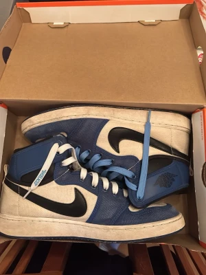 Nike Jordan 1 blå och vita sneakers - Säljer ett par Nike Jordan 1 sneakers i blått och vitt med svarta swoosh-detaljer. Skorna har högt skaft, rund tå och snörning med både vita och blå skosnören. Materialet är en mix av canvas och syntet, och de har klassisk Jordan-logga på sidan.