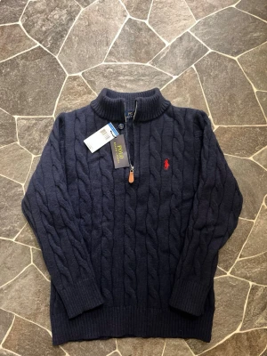 Marinblå stickad tröja Polo Ralph Lauren - Säljer en marinblå stickad tröja från Polo Ralph Lauren med kabelstickat mönster och röd broderad logga på bröstet. Tröjan har half zip med läderdetalj, ribbad krage och långa ärmar. Perfekt för kyliga dagar och snygg till jeans. Passar nog M - S!