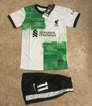 Liverpool fotbollströja och shorts Nike - Liverpool matchställ med M. Salah och nummer 11. Tröjan är vit med gröna grafiska detaljer, svarta muddar och klubbmärke på bröstet. Svarta shorts med vit rand och nummer 11. Perfekt för fotbollsfans som vill ha en cool look på planen.