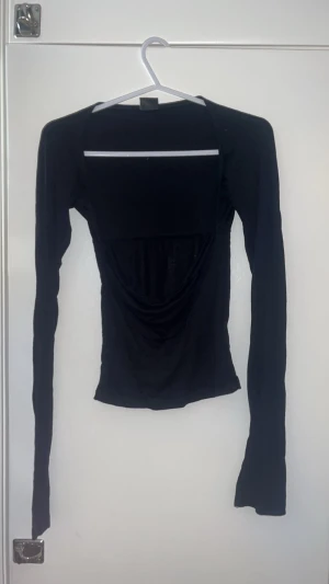 Svart bolerotopp med drapering - Säljer en svart bolerotopp med lång ärm och snygg drapering framtill. Toppen har en smal passform och är tillverkad i ett mjukt, stretchigt material som sitter skönt mot kroppen. Perfekt att styla med höga jeans eller kjol för en cool look.