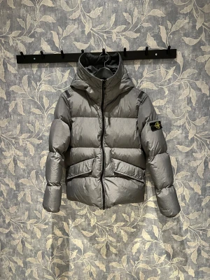 Grå pufferjacka från Stone Island - Snygg grå pufferjacka från Stone Island Junior med huva och ikonisk patch på ärmen. Jackan har två stora fickor framtill, dragkedja och en puffig siluett som ger en cool vibe. Perfekt för kalla dagar när du vill hålla stilen. Passform loose