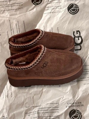 Ugg 39 nya  - Säljer ett par nya bruna slip-on skor från UGG med mjukt pälsfoder och mönstrad kant i beige och brunt. Skorna har en platt sula och är tillverkade i mocka med UGG-logga på sidan. Perfekta för chill dagar och har en cozy vibe.