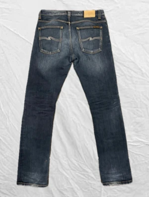Nudie jeans mörkblå - Snygga Nudie jeans i mörkblå tvätt med klassiska fem fickor och gul kontrastsöm. Modellen har rak passform och normal midja, med Nudie-logga på bakfickan och läderpatch i midjan. Perfekta för dig som gillar stilrena jeans med lite attityd. Storlek: W30 & L32. Modell: GRIM TIM