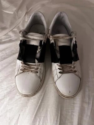 Valentino Garavani vita sneakers - Säljer ett par vita sneakers från Valentino Garavani med bred svart detalj över sidan. Skorna har snörning och rund tå, tillverkade i skinn med platt sula. Klassisk och stilren design som passar till många olika outfits. Pris kan diskuteras 