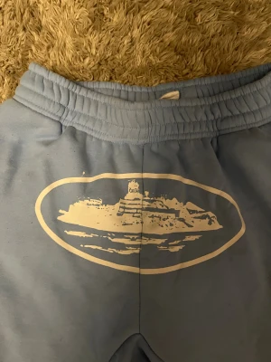 Blåa corteiz alcatraz shorts - Säljer ett par ljusblå cortiez mjukisshorts i storlek M med elastisk midja och vit snörning. Framtill finns ett stort vitt tryck med motiv av en byggnaden alcatraz. Shortsen har fickor på sidorna och en bakficka med knapp. Perfekta för chill dagar.