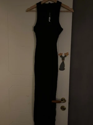 Svart långklänning  - Snygg svart långklänning med ärmlös design och rund hals. Klänningen har en slim passform som framhäver figuren och är perfekt för dig som gillar stilrena och enkla plagg. Materialet känns mjukt och följsamt mot kroppen.