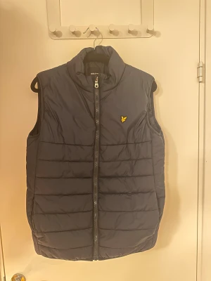 Mörkblå dunväst från Lyle & Scott - Snygg mörkblå dunväst från Lyle & Scott Junior med gul broderad logga på bröstet. Västen har quiltad design, hög krage och dragkedja framtill. Perfekt att slänga på över en hoodie för extra värme. Passar dig som gillar stilren och sportig look.