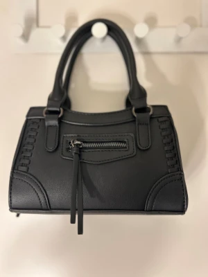 Svart handväska med axelrem - Snygg svart handväska i skinnimitation med både handtag och justerbar axelrem. Väskan har dekorativa sömmar och en liten dragkedja framtill med lång rem. Perfekt storlek för det viktigaste och stilren design som passar till många outfits.