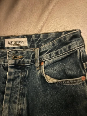 Blå jeans från Just Junkies - Säljer ett par klassiska blå jeans från Just Junkies med rak passform och ganska högmidjad. Den sluyar precis under naveln. Jeansen har fem fickor, knappgylf och är tillverkade i robust denim. 