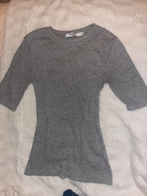 Grå ribbad topp från NA-KD - Säljer en enkel och stilren grå ribbad topp från NA-KD i storlek XS. Toppen har rund halsringning och korta ärmar, med en slim passform som sitter snyggt på kroppen. Perfekt att matcha med jeans eller kjol för en clean look.