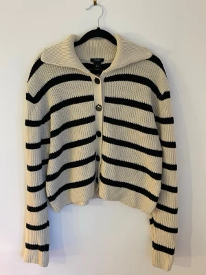 Randig kofta från Lindex  - Säljer en snygg stickad kofta från Lindex i storlek S. Koftan är beige med svarta ränder, har stora knappar framtill och en bred krage. 