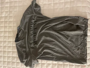 Grå t-shirt från Emporio Armani - Snygg grå t-shirt från Emporio Armani med svart tryck på framsidan. Klassisk rund hals och korta ärmar. Materialet känns mjukt och lätt, perfekt för en chill stil. Passar dig som gillar stilrena märkesplagg med diskret logga.