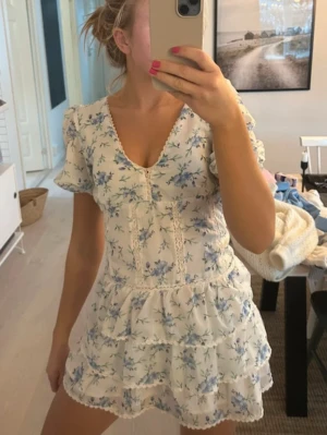 Vit blommig kortklänning med volanger - Superfin vit kortklänning med blå blommigt mönster och volanger. Klänningen har v-ringad hals, puffiga korta ärmar och spetsdetaljer längs sömmarna. Perfekt för sommarens alla tillfällen, riktigt söt och romantisk stil.