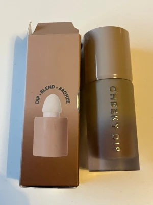 Cheeky Dip Liquid Bronzer - Flytande bronzer från Cheeky Dip i en snygg beige-brun förpackning med inbyggd applikator. Ger en naturligt solkysst look och är enkel att blanda ut tack vare den mjuka svampapplikatorn. Perfekt för att framhäva ansiktets konturer med en varm ton.