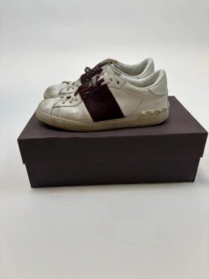 Valentino Open sneakers i vitt och mörkröd - Säljer ett par Valentino Open sneakers med vit skinnbas och bred mörkröd panel på sidan. Skorna har snörning och platt sula med ikoniska nitar vid hälen. Perfekt för dig som gillar lyxiga detaljer och stilren design.