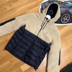 Beige teddyjacka från Polo Ralph Lauren - Snygg teddyjacka från Polo Ralph Lauren i beige och svart. Jackan har huva med dragsko, dragkedja framtill och två fickor med dragkedja. Överdel och ärmar i mjuk teddy, nederdel i quiltad svart polyester. Perfekt för kalla dagar och riktigt stilren look.