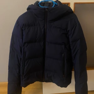 Mörkblå vinter pufferjacka med huva - Säljer en mörkblå  vinter- pufferjacka med huva och dragkedja framtill. Jackan har två stora fickor med dragkedja på utsidan och två innerficka med knapp. Perfekt för kalla dagar och riktigt skön att bära. Storlek XS men passar S 
