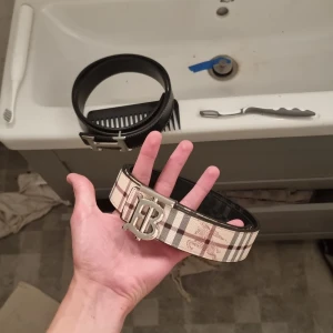 Burberry bälte  - Snyggt bälte med klassiskt Burberry-rutigt mönster i beige, brunt och svart. Bältet har en stor silvrig metallspänne med ett B framtill. Perfekt accessoar för att lyfta din outfit med en lyxig touch.