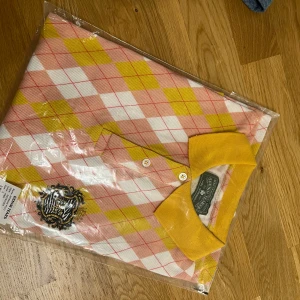 Rutig pikétröja från Denim Tears - Säljer en riktigt snygg pikétröja från Denim Tears med rutigt mönster i rosa, gult och vitt. Tröjan har en gul krage, knappar framtill och en broderad patch med zebramönster och gulddetaljer på bröstet. Perfekt för dig som vill sticka ut med färg och mönster.