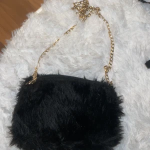 Svart fluffig axelväska med guldkedja - En svart fluffig axelväska med mjuk pälsimitation och snygg guldfärgad kedja som axelrem. Väskan har dragkedja upptill och ett rymligt fack inuti. Perfekt för dig som vill sticka ut med en cool accessoar.