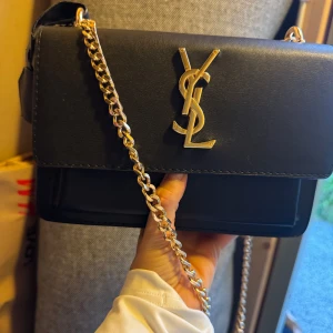 Svart YSL axelväska med guldkedja - Snygg svart axelväska från YSL med ikoniskt guldigt logospänne framtill och en chunky guldfärgad kedja som axelrem. Stilren och lyxig design i slätt skinn, perfekt för dig som vill ha en statementväska med exklusiv känsla.helt ny