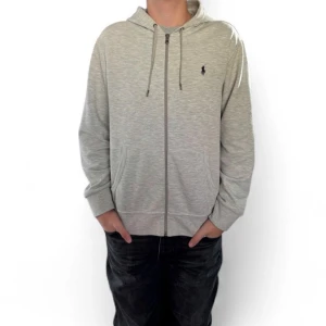Grå hoodie från Polo Ralph Lauren - Snygg ljusgrå hoodie från Polo Ralph Lauren med dragkedja framtill, huva med snörning och två fickor. Klassisk logga broderad på bröstet. Tillverkad i mjuk bomullsmix, perfekt för chill dagar eller när du vill ha en clean look.