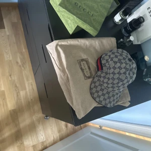 Gucci keps med GG-mönster och läder - Snygg keps från Gucci med klassiskt GG-monogram i blått på beige bakgrund. Kepsen har justerbar rem i svart läder och detaljer med röd och marinblå rand på sidan. Materialet är bomull och läder, perfekt för dig som vill ha en lyxig streetstyle-accessoar.