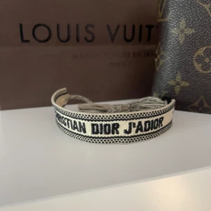 Vit och svart Dior armband - Snyggt beige och svart armband från Christian Dior med tydlig logga framtill. Armbandet är vävt i bomull och har svarta ränder samt knytning med fransar baktill. Perfekt för dig som gillar designeraccessoarer med cool vibe.