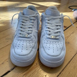 Nike Air Force 1 vita sneakers - Fräscha Nike Air Force 1 sneakers i helvitt skinn med klassisk låg siluett. Snygga perforeringar på tån, tydlig swoosh på sidan och AIR-logga bak. Platt sula och snörning framtill. Perfekta för dig som gillar clean och tidlös streetstil.