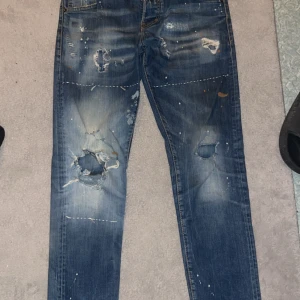 Dsquared2 jeans - Riktigt snygga blå jeans från Dsquared2 med slitningar, hål och färgstänk för en rå streetlook. Jeansen har klassisk femficksdesign, smal passform och coola detaljer som ger dem en unik vibe. Perfekta för dig som gillar statement-plagg. Pris kan diskuteras 