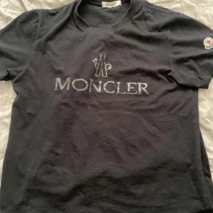 Svart Moncler t-shirt med logga - Svart t-shirt från Moncler med stor logga tryckt på bröstet och klassisk rund hals. På ärmen sitter en broderad Moncler-patch i vitt, blått och rött. T-shirten är i mjuk bomull och har en normal passform. Perfekt för dig som gillar stilrena och exklusiva plagg.