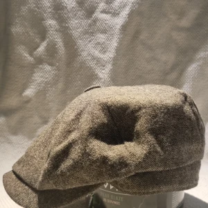 Grå newsboy keps från Heritage Traditions - Snygg newsboy keps i beige från Heritage Traditions. Kepsen har klassisk form med mjuk, rundad skärm och är tillverkad i grovvävd bomullsliknande textil. Insidan är quiltad för extra komfort. Perfekt för dig som gillar vintagevibbar och tidlös stil.